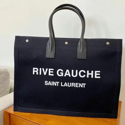 A navy canvas Saint Laurent Rive Gauche tote with black leather handles, standing on a wooden table
